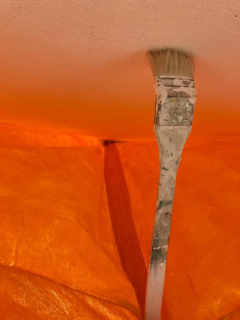 The artist Francis Ruyter has taken a photo of a paintbrush which is holding up a tent. The brush is suspended between a white surface and an orange-colored canvas tent. The white surface reflects the orange color.